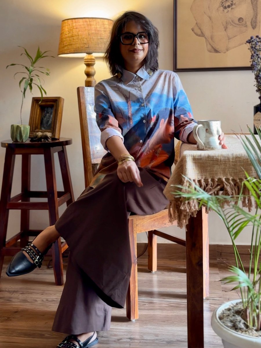 Woman wearing the Shakuntala shirt with brown trousers, seated on a wooden chair, full length view